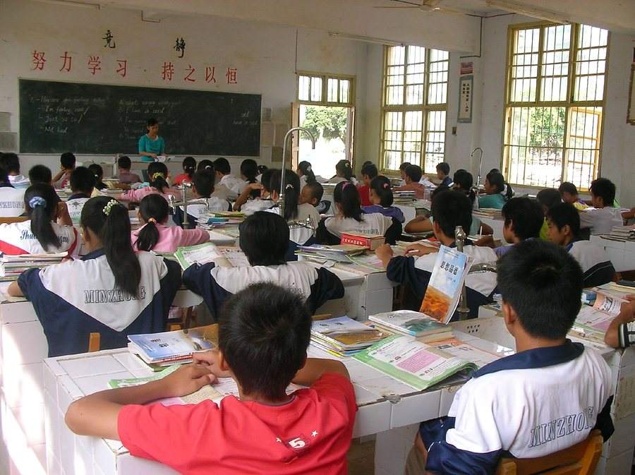 A full classroom of students in uniform reading at their desks, their teacher at the blackboard