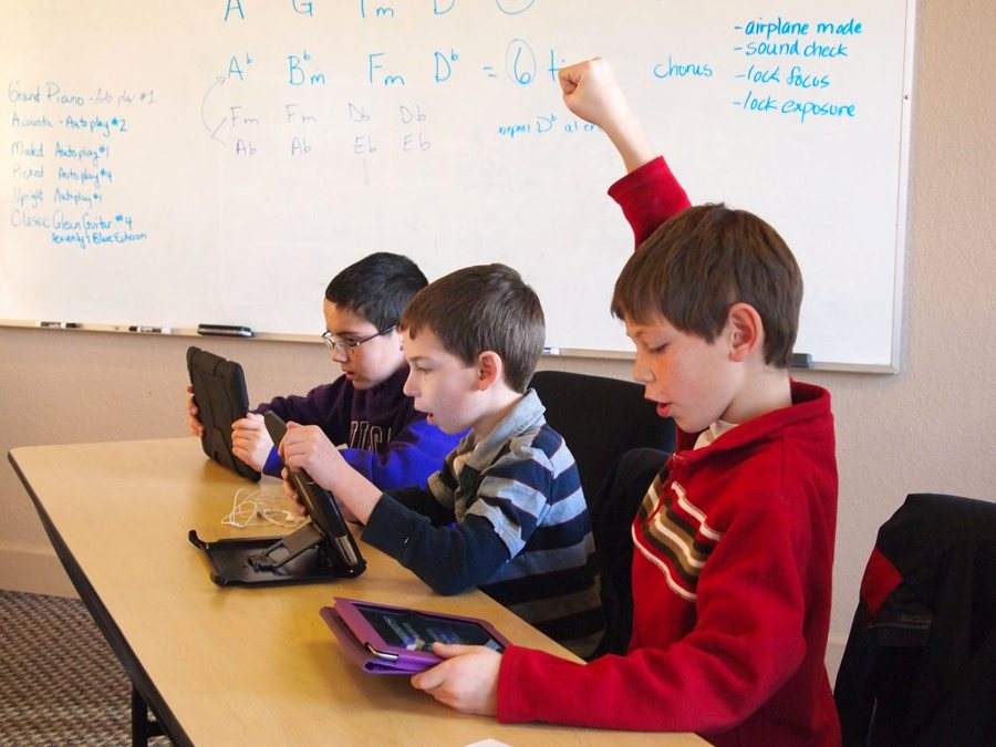 Three boys working on tablets at a classroom table, one with his hand raised