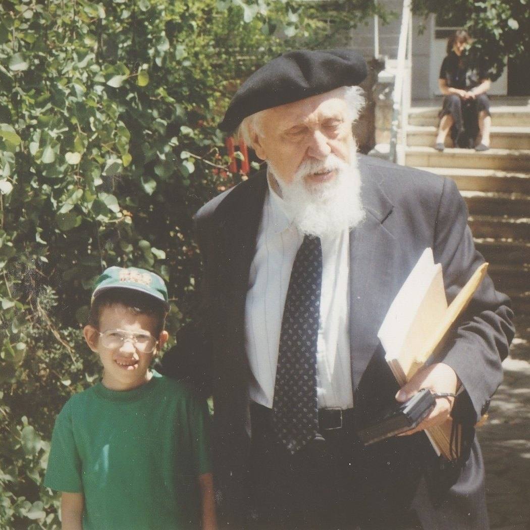 Professor Reuven Feuerstein in a beret and suit, talking in a sunlit garden beside a smiling young boy in a cap and glasses